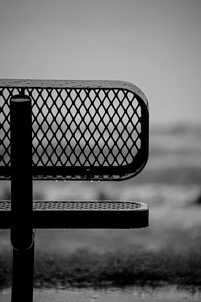 An example of black and white photography that shows a metal park bench with raindrops clinging to the lower edges of the seat and backrest.
