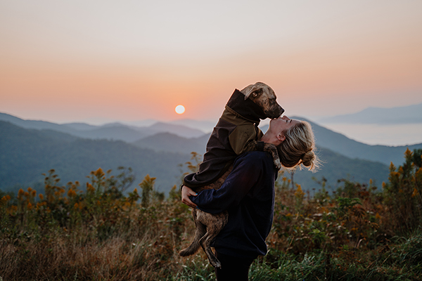 Woman posing holding and kissing dog wearing jacket in front of sunset